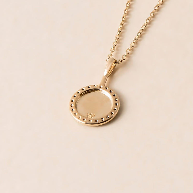 The back of a gold pendant necklace from the Lua Collection, featuring a circular gold pendant with an intricate engraving, displayed on a light beige background.