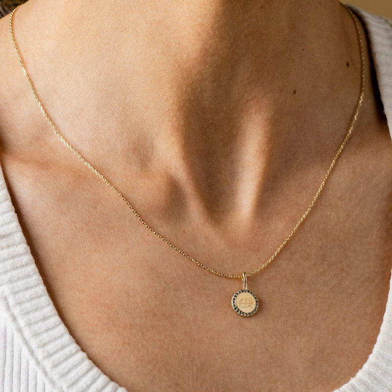 a person wearing a gold necklace with a round pendant featuring an engraved greatest name symbol, encircled by blue diamonds..