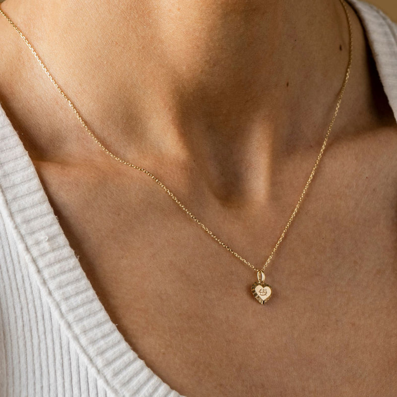 A person wearing The Golden Love Heart necklace - a gold heart-shaped pendant with a decorative edge, engraved Greatest Name, complemented by radiant engraved sunrays.