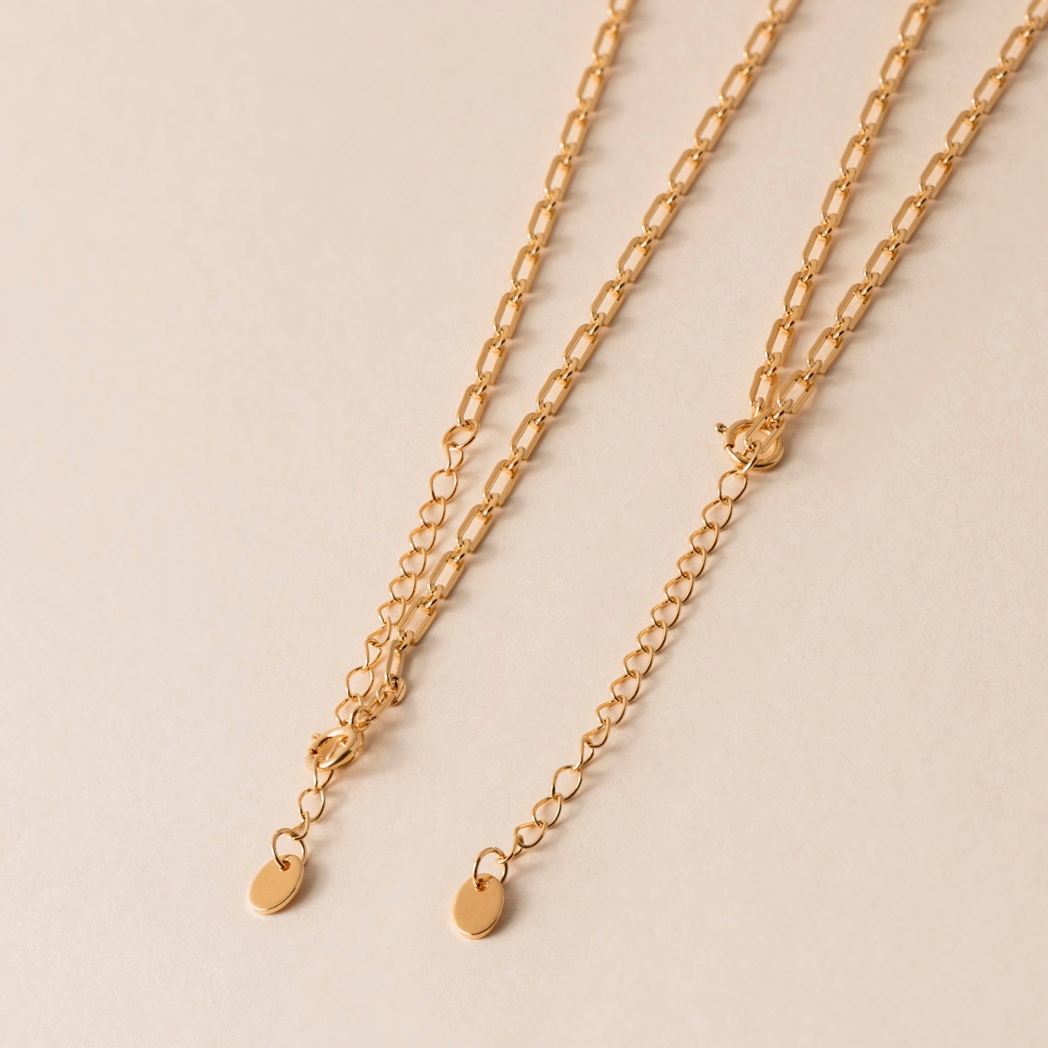 Close-up of two 18k Gold Vermeil chains with oval links and small oval lua tags at the ends, beautifully displayed on a light beige background. 