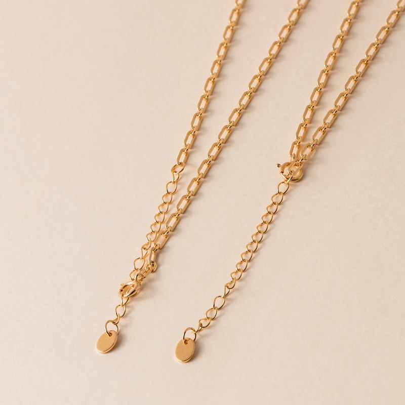 Close-up of two 18k Gold Vermeil chains with oval links and small oval lua tags at the ends, beautifully displayed on a light beige background. 