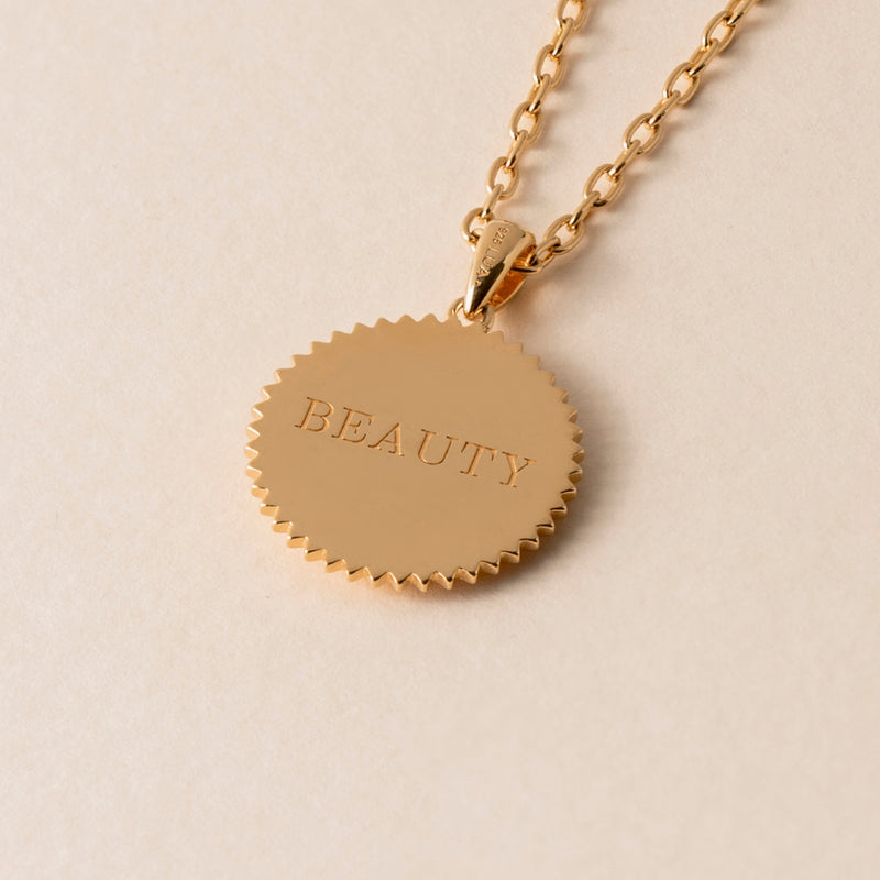 A gold necklace with a circular pendant that has the word "BEAUTY" engraved on it is displayed against a light beige background, showcasing luxurious textures.