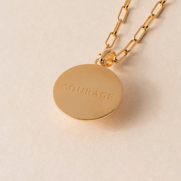 A gold necklace with a circular pendant engraved with the word "COURAGE," made from 18k Gold Vermeil, is displayed on a neutral background.