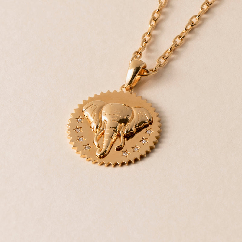 Close-up of an Elephant Necklace featuring a circular pendant with an embossed elephant head and five small star engravings, symbolizing compassion. The gold chain has an interlocking design. The background is a plain beige surface.