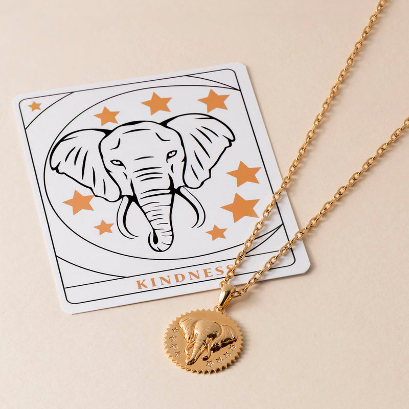 A gold Kindness Elephant Necklace with an elephant pendant lies beside a card showcasing an elephant graphic and the word "KINDNESS" at the bottom. Both items are adorned with star illustrations, symbolizing compassion and elegance.