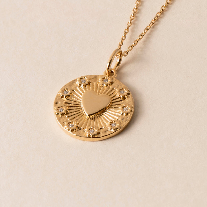 The Love Heart Necklace is a gold necklace with a round pendant featuring a raised heart in the center, surrounded by diamond-like studs and radiating lines. This thin, delicate chain symbolizes pure love, making it an elegant symbol of affection.