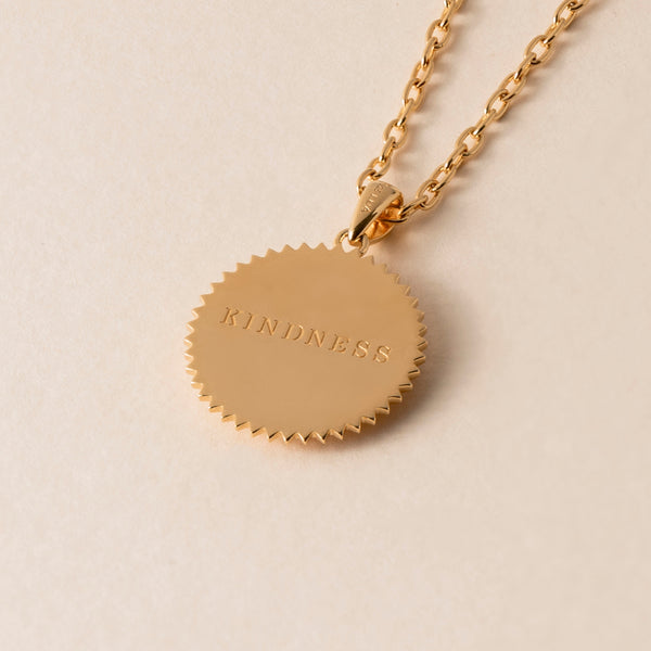 A Kindness Elephant Necklace featuring a gold chain and a round, jagged-edged pendant engraved with the word "KINDNESS." The elephant symbolizes a compassionate nature, making this piece both meaningful and elegant against the plain, beige background.