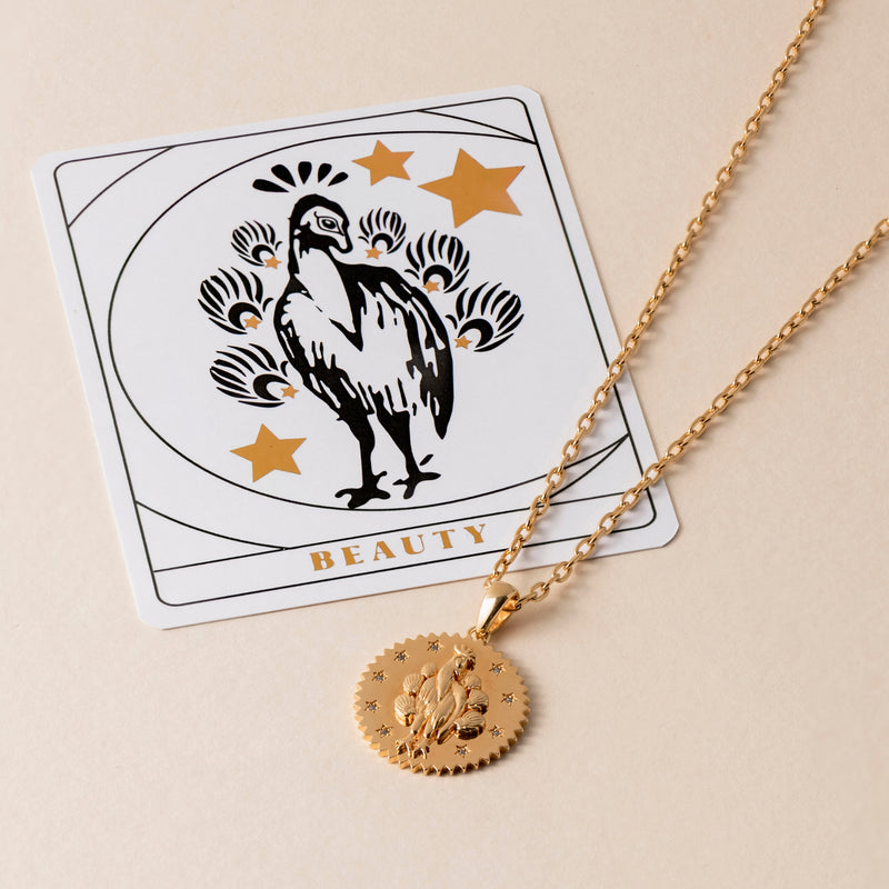 A Peacock Necklace with a round pendant featuring a bird design lies over a beauty-themed card with peacock and star illustrations.