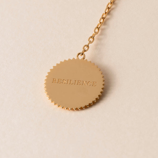 A gold pendant with the word "RESILIENCE" engraved on it hangs from a delicate gold chain, embodying the spirit of transformation like a butterfly necklace, set against a beige background.