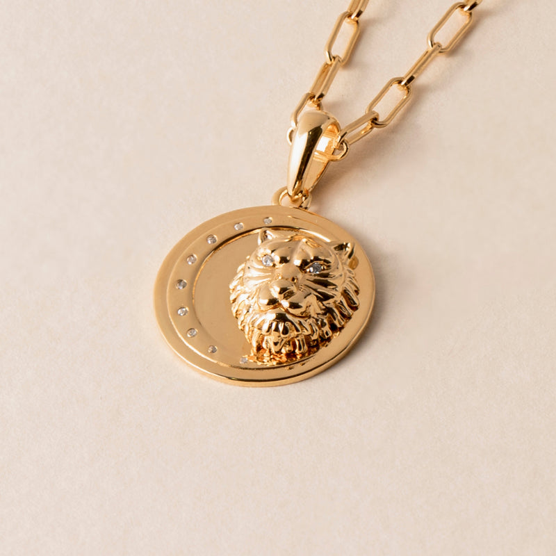 A gold necklace with a pendant depicting a lion's head, adorned with small gems around the top half of the circular border, is crafted in 18k Gold Vermeil and suspended from a chain of adjustable length.