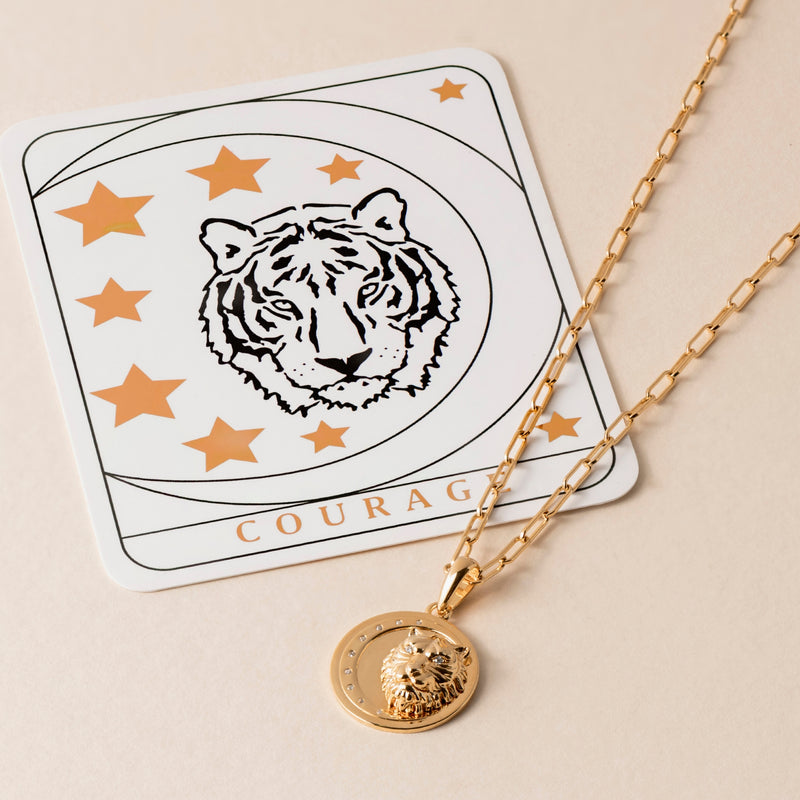 A gold necklace with a pendant featuring a lion's head lies next to a card displaying a tiger illustration and the word "Courage." Crafted from 18k gold vermeil, this piece is both elegant and symbolic.