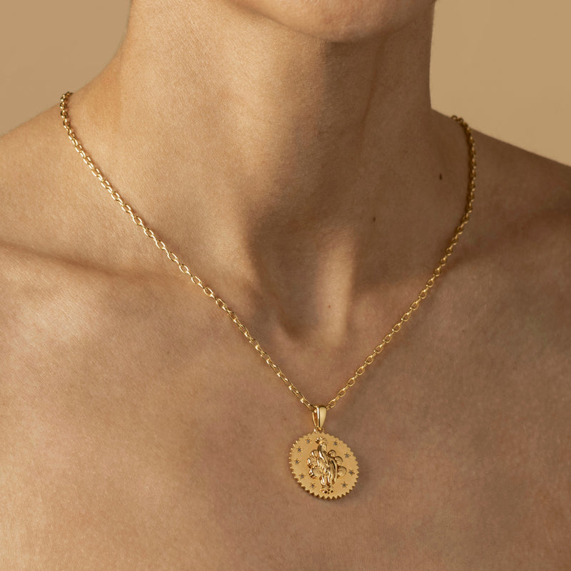 A person wearing a gold necklace with a round, intricately embossed peacock pendant on a light brown background experiences the beauty of this delicate adornment.