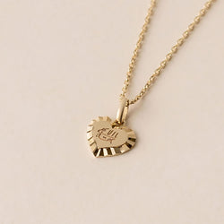 The Golden Love Heart necklace features a gold heart-shaped pendant with a decorative edge, hanging from a gold chain. It showcases engraved Greatest Name, complemented by radiant engraved sunrays.