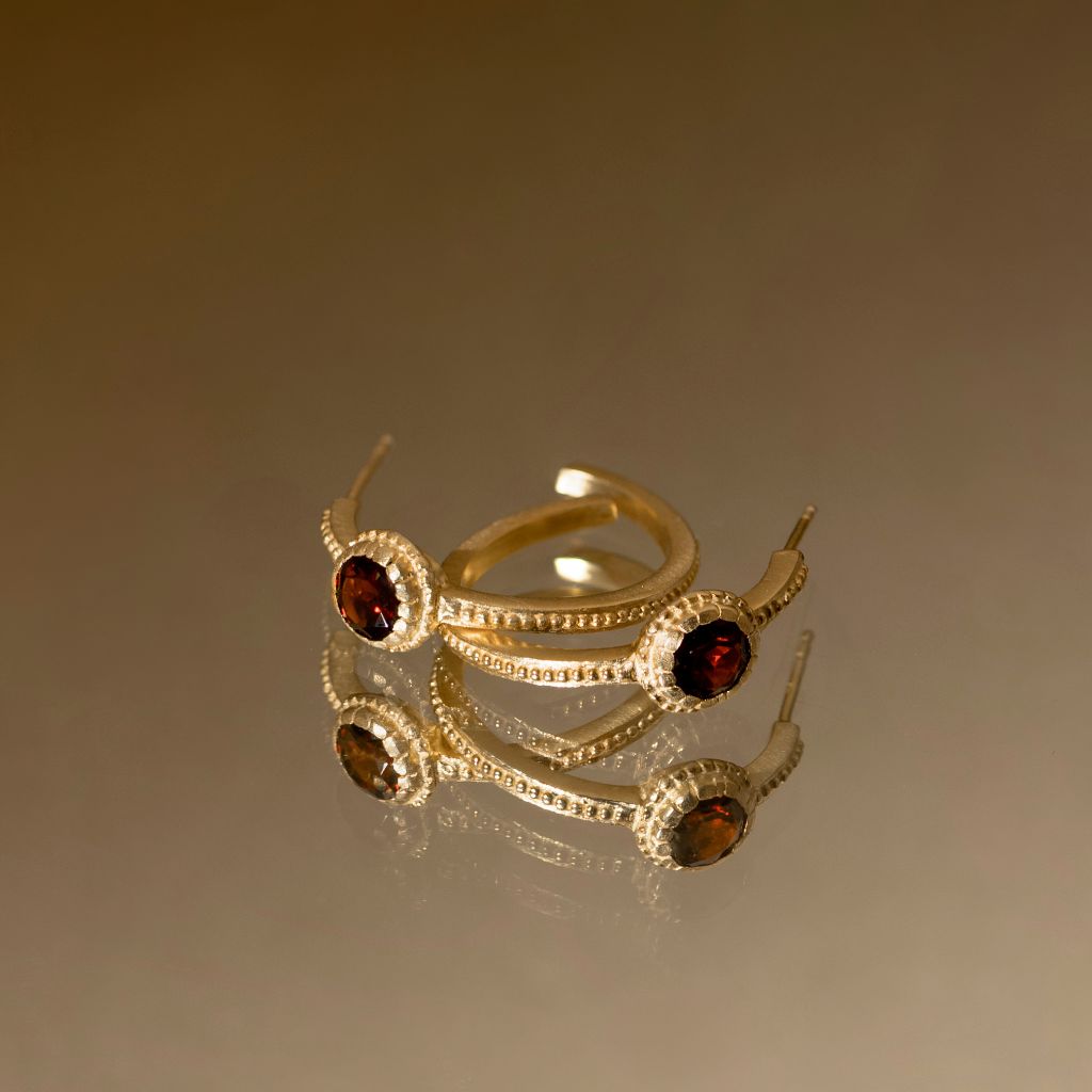 A gold-plated open ring band featuring three circular settings with dark red garnet-like stones and small clear crystals, displayed on a reflective surface against a warm brown gradient background.