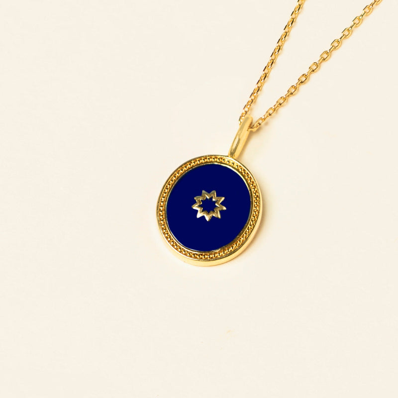 Gold necklace with a blue enamel pendant featuring a nine-pointed star-shaped cutout, displayed against a plain beige background.