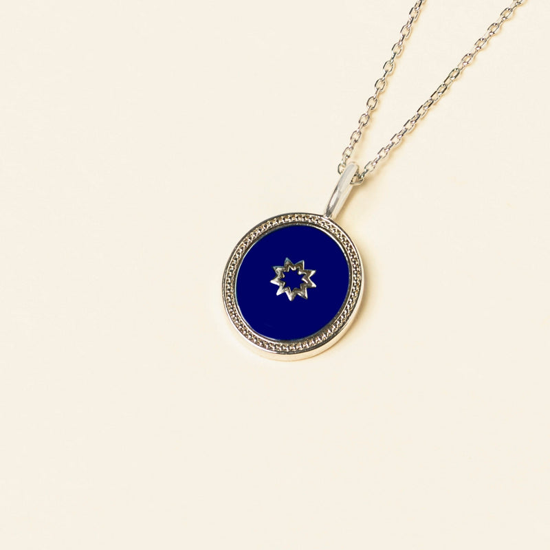 A circular blue pendant featuring the Greatest Name Symbol, surrounded by a silver frame, hangs on a chain against a pale background.