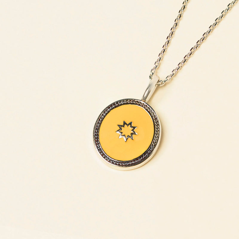A pendant with a bespoke enamel center featuring a small starburst design, surrounded by a silver frame, on a silver chain against a cream background.