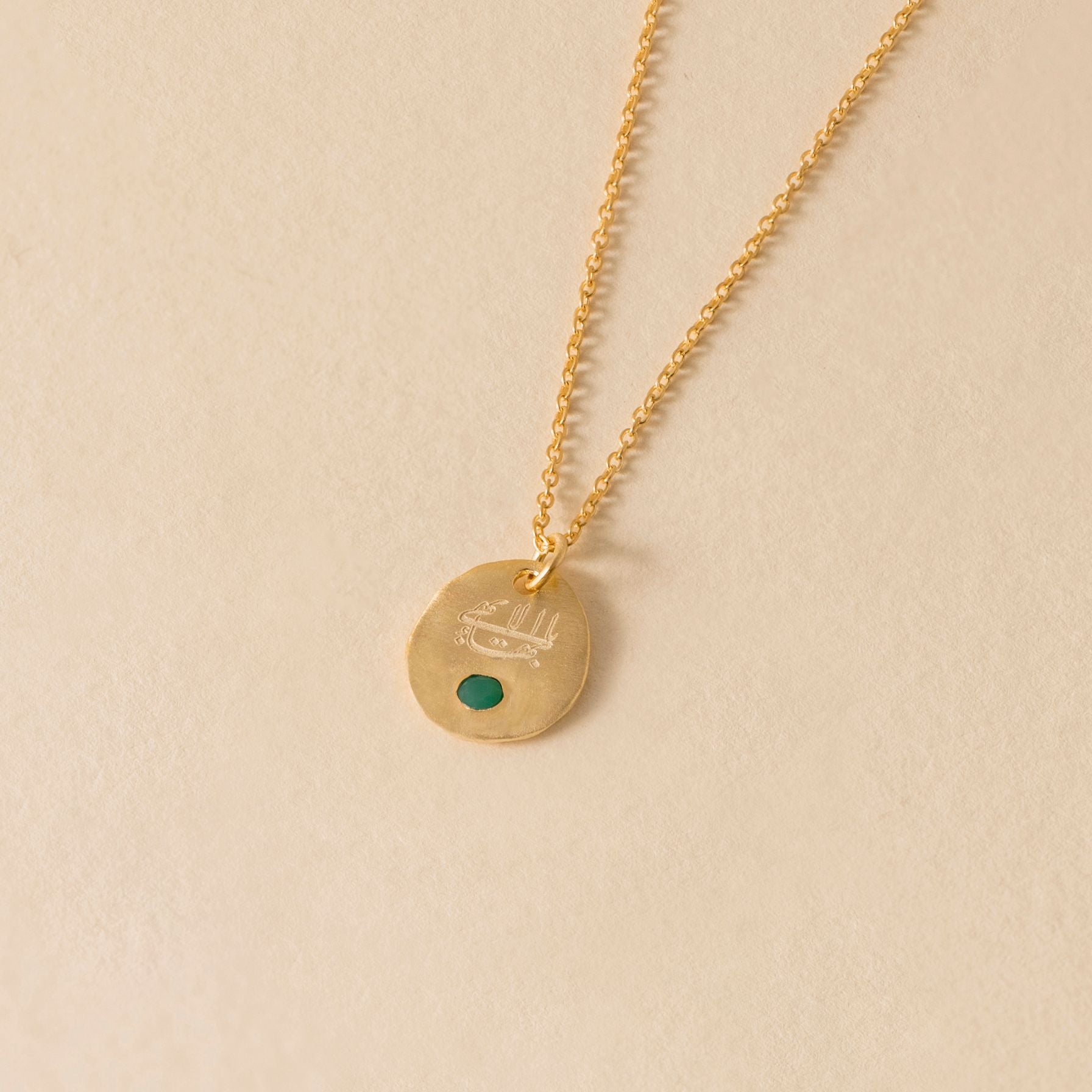 Gold necklace with an oval pendant featuring an emerald stone and the greatest name on a beige background
