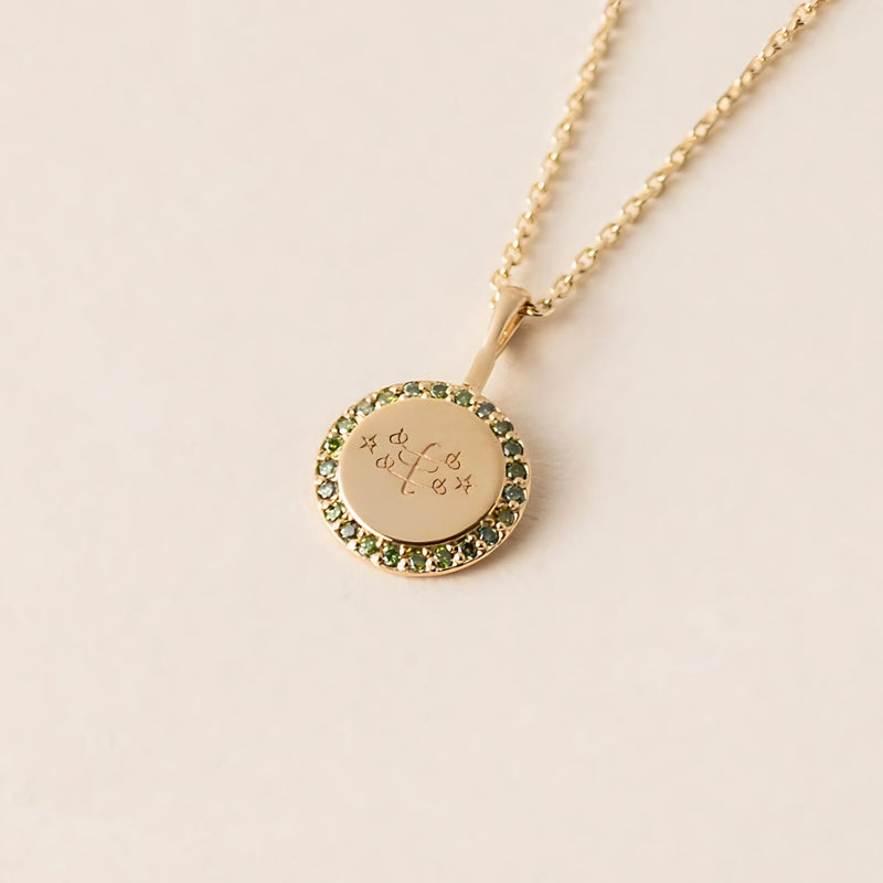 A gold necklace with a round pendant featuring an engraved ringstone symbol, encircled by green diamonds, displayed against a neutral background.