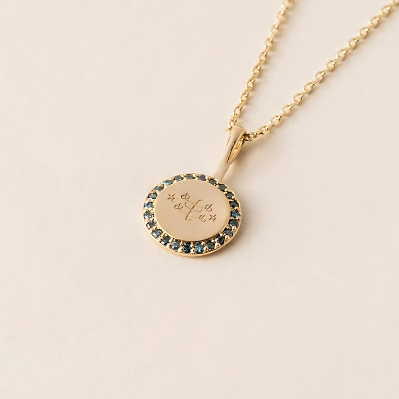 A gold necklace with a round pendant featuring an engraved Ringstone symbol, encircled by blue diamonds, on a cream background.