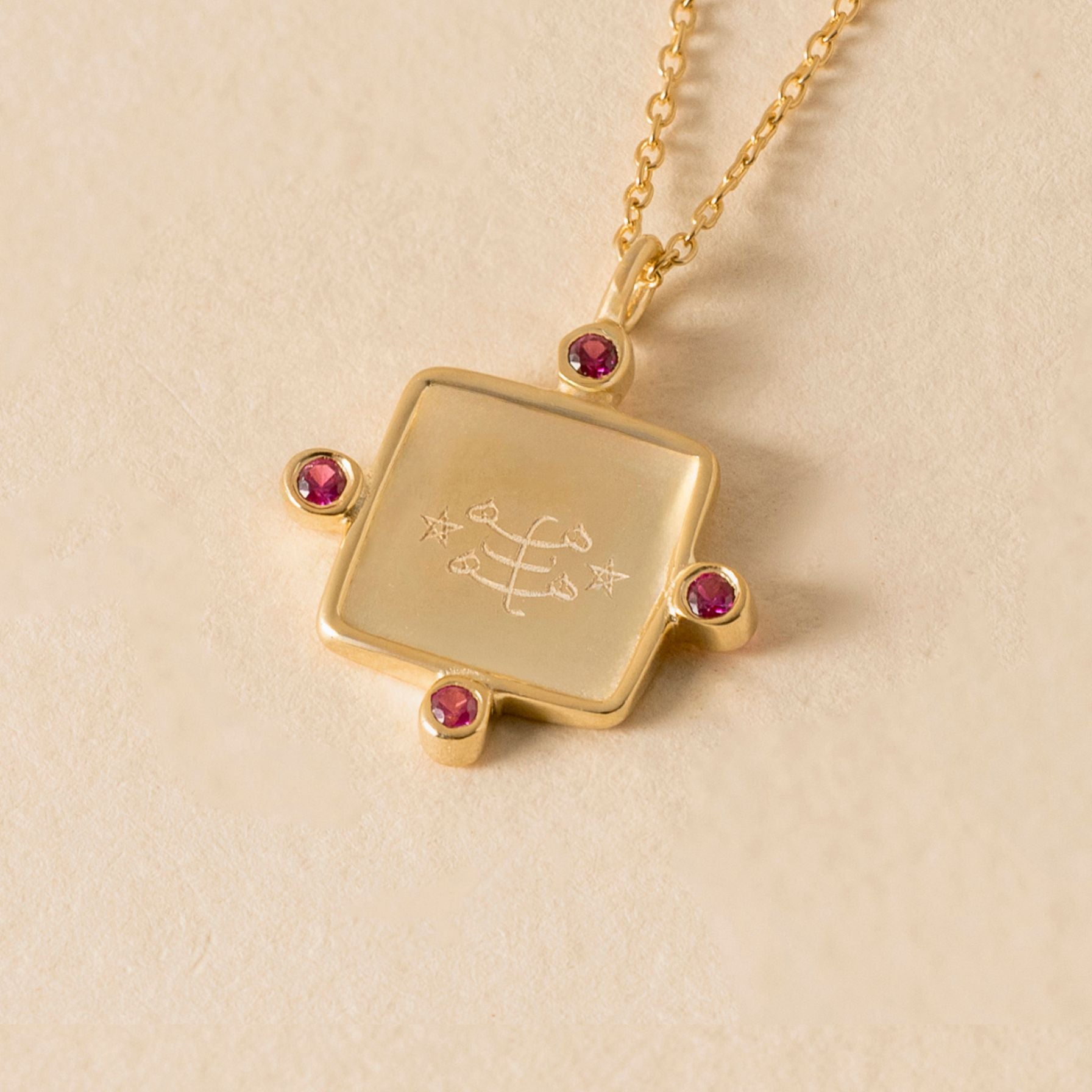 Gold necklace with a square pendant featuring ringstone symbol engraved design and pink gemstones on a beige background