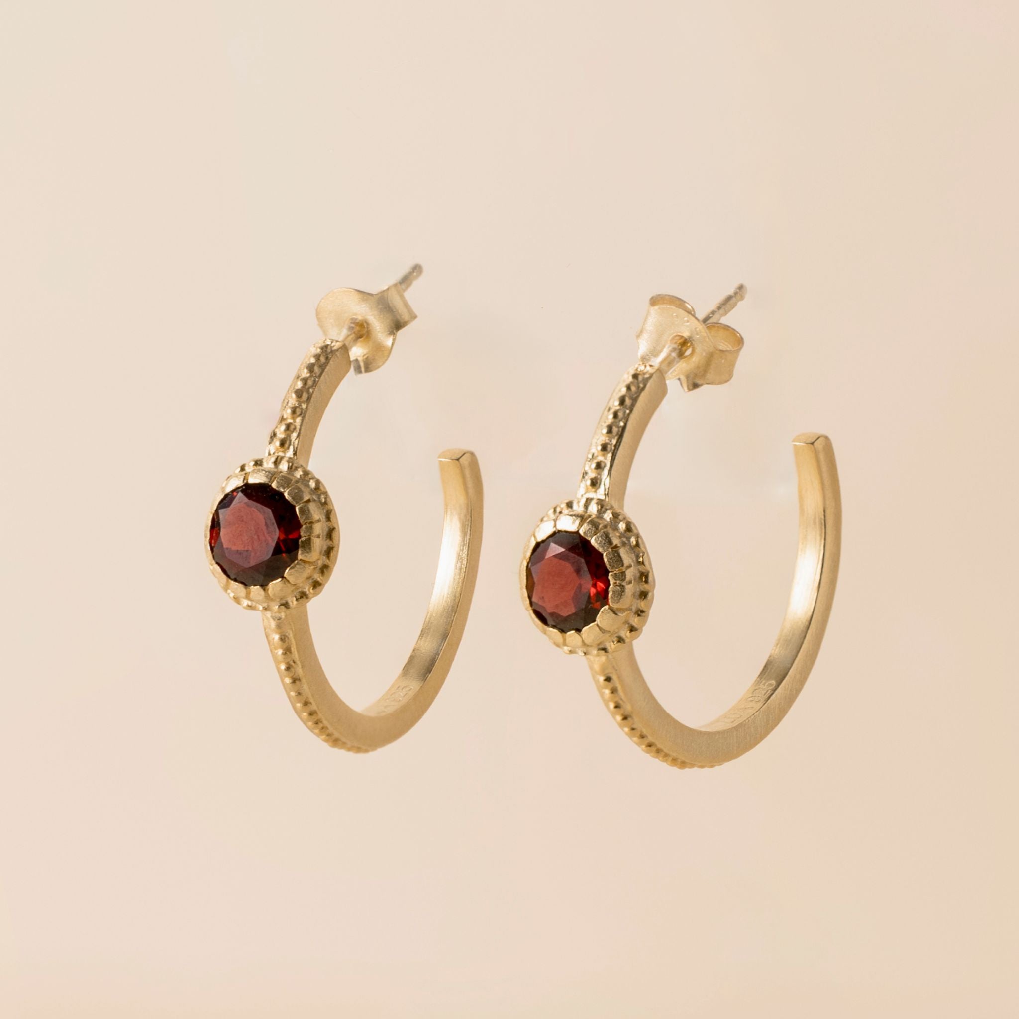 A pair of gold huggie hoop earrings with textured beaded borders and round dark red gemstone centers, displayed on a solid light beige background.