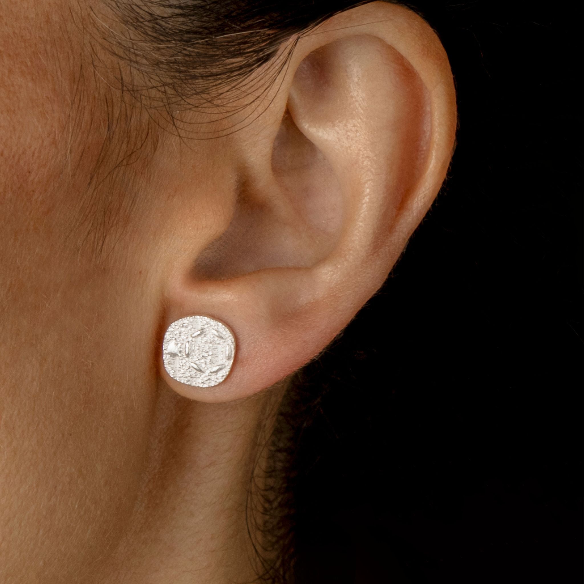 A close-up of a person's ear wearing a round, textured silver stud earring against a solid black background.