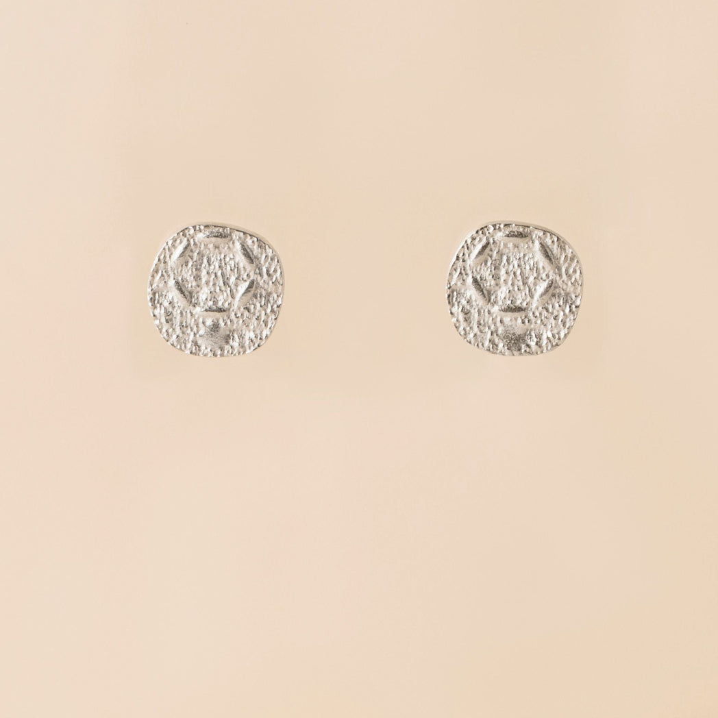 A pair of textured silver stud earrings with a circular, medallion-like design, displayed on a solid beige background.