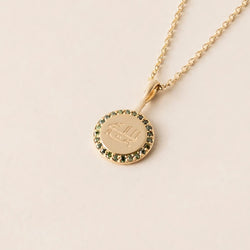 A gold necklace with a round pendant featuring an engraved greatest name symbol, encircled by green diamonds, displayed against a neutral background.