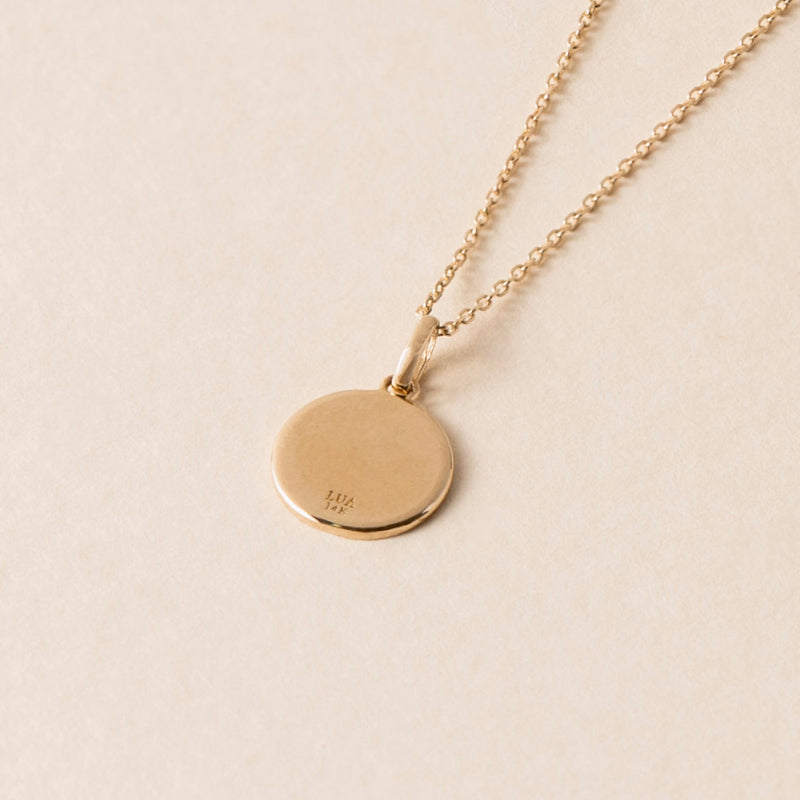 The back of a gold pendant necklace from the Lua Collection, featuring a circular gold pendant with an intricate engraving, displayed on a light beige background.